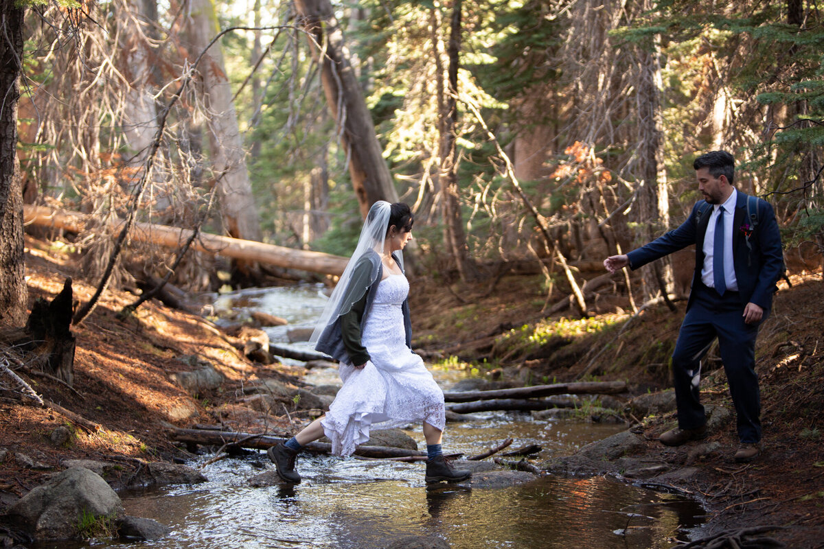 A groom stands on one side of a creek holding out his hand to his bride who jumps across.