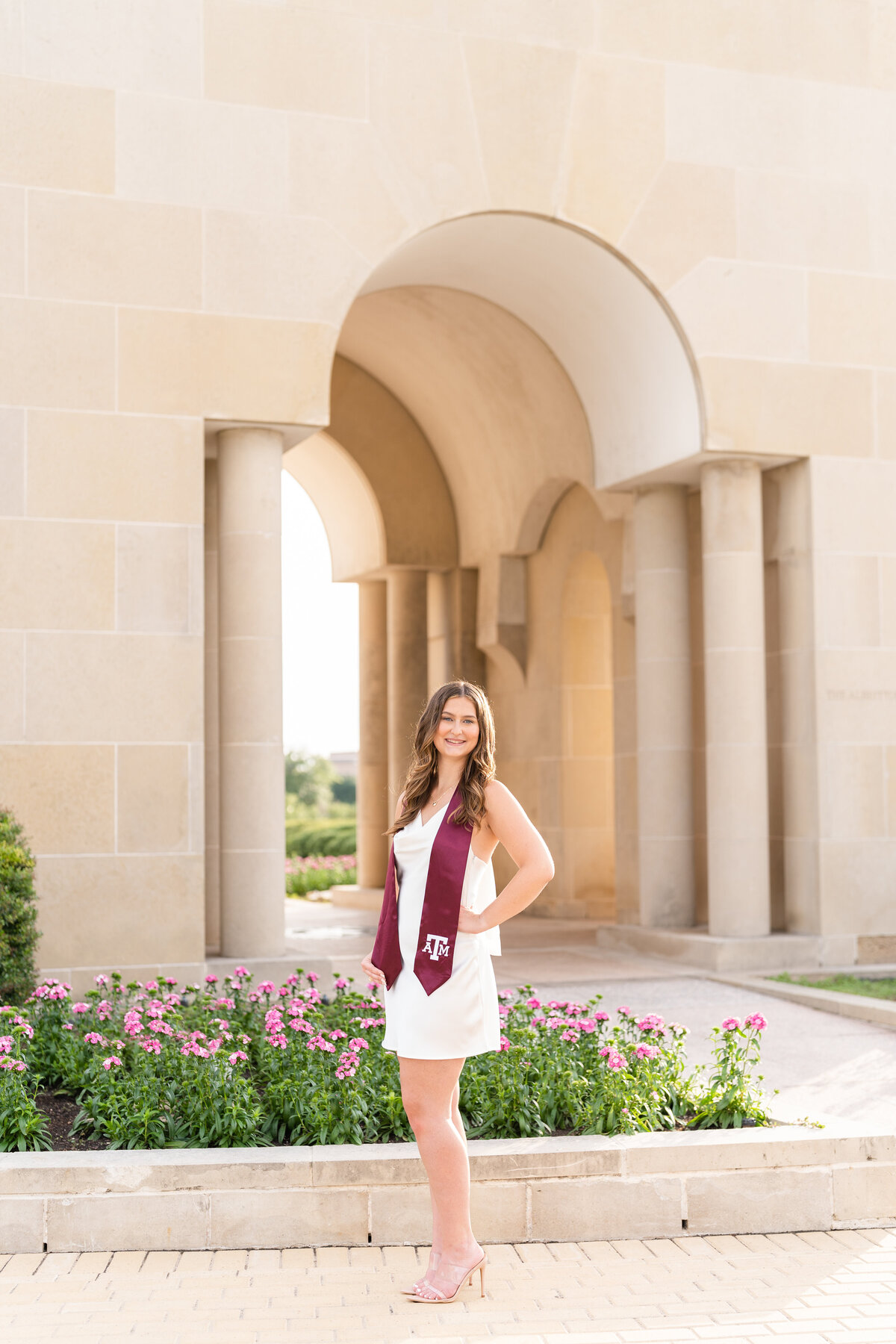 Texas A&M senior girl standing and smiling in front of Bell Tower while wearing white dress and maroon stole with hand on hip