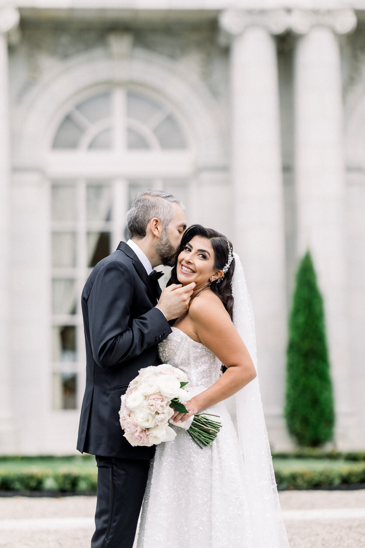 sophisticated bride and groom  kiss outside during wedding day portraits outside of rosecliff mansion in newport rhode island, groom kisses her on the temple during first look