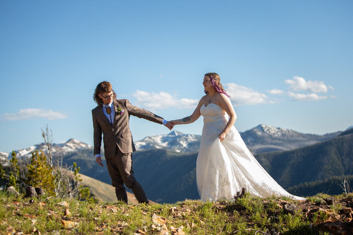 A groom leads his bride across a grassy hill with mountains in the background.