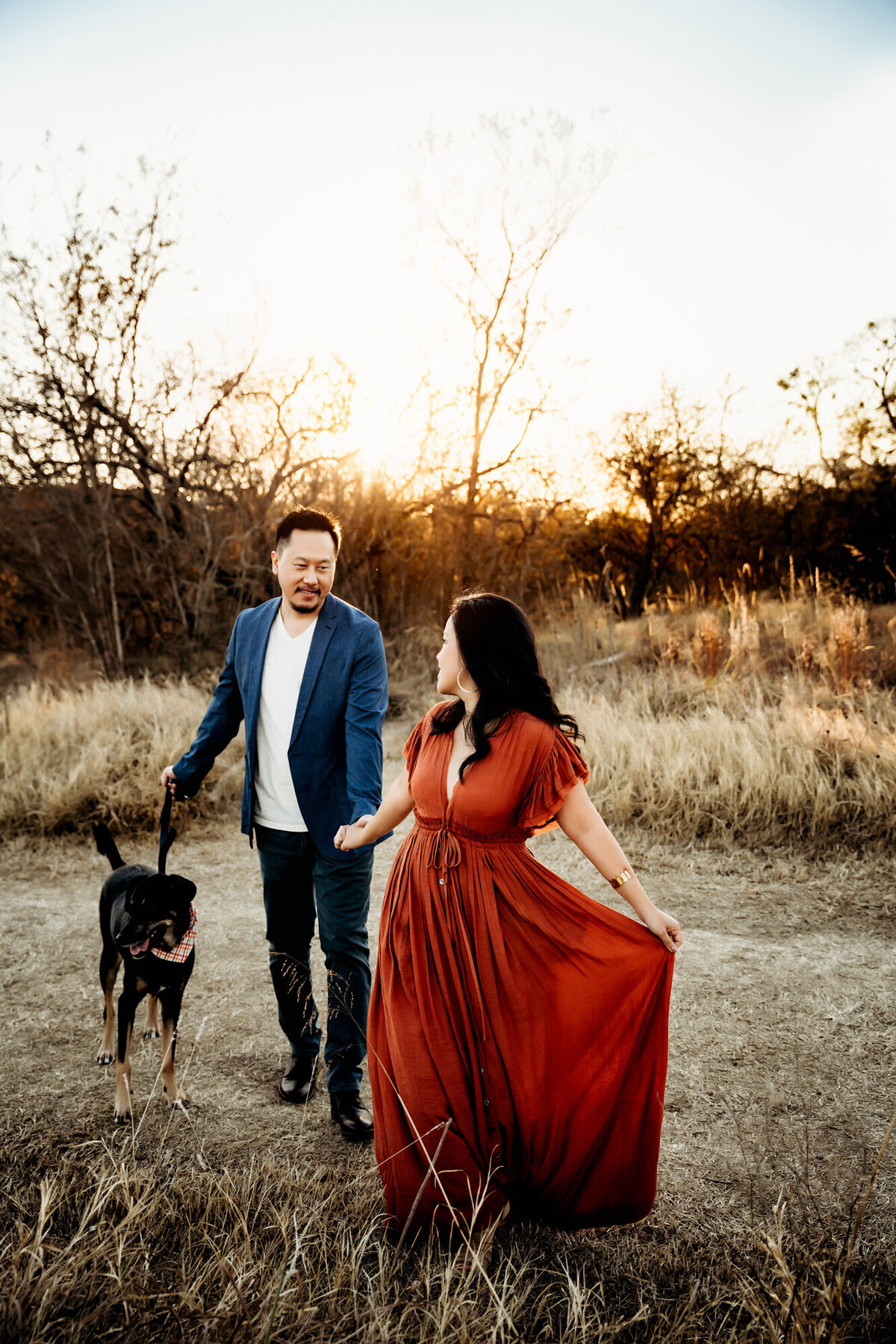 Couples Photographer,  Woman in a rust colored dress walking in a field holding hands with a man in a blue blazer who is walking a dog. This is at sunset.