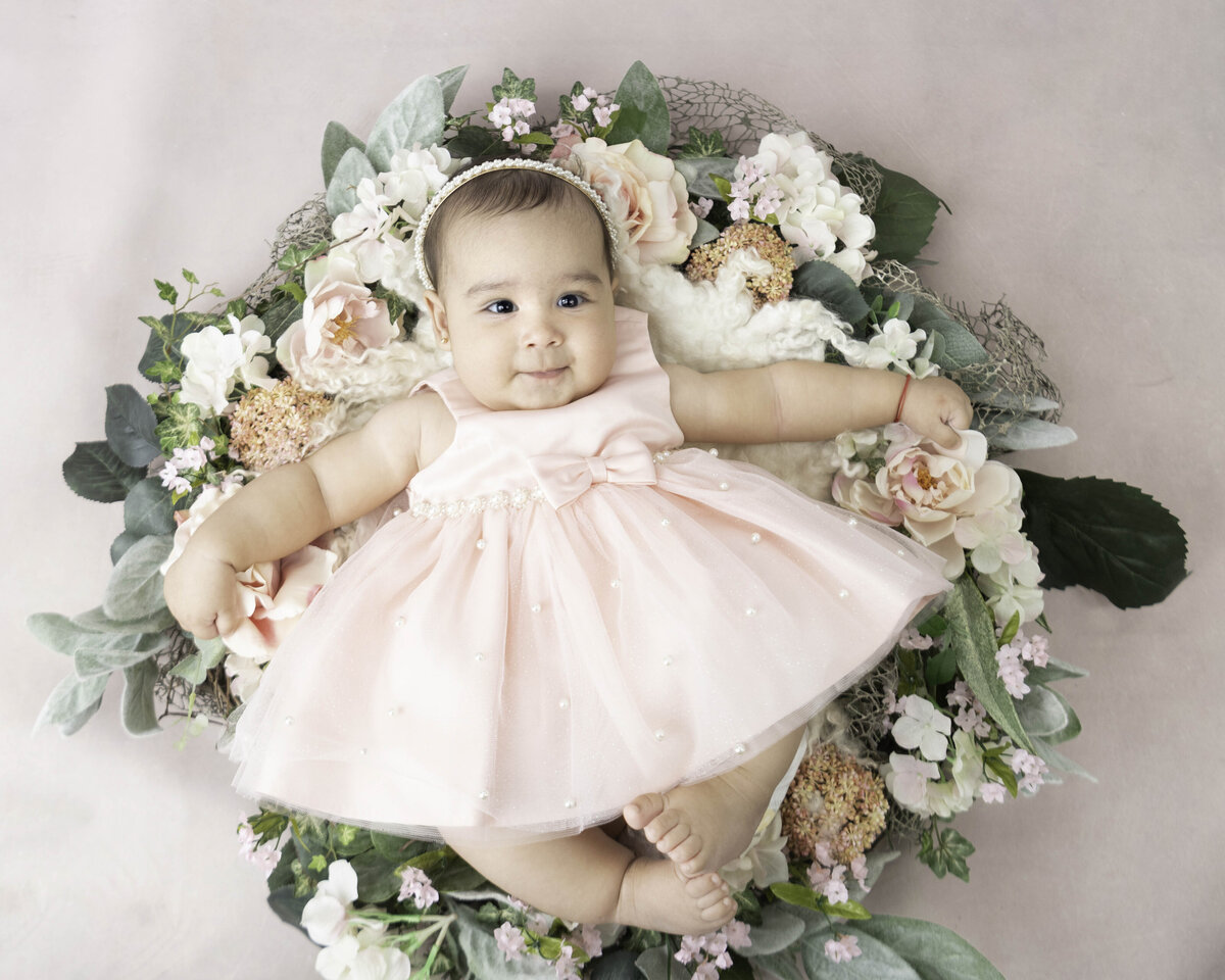A happy baby lays on a bed of flowers in a pink dress