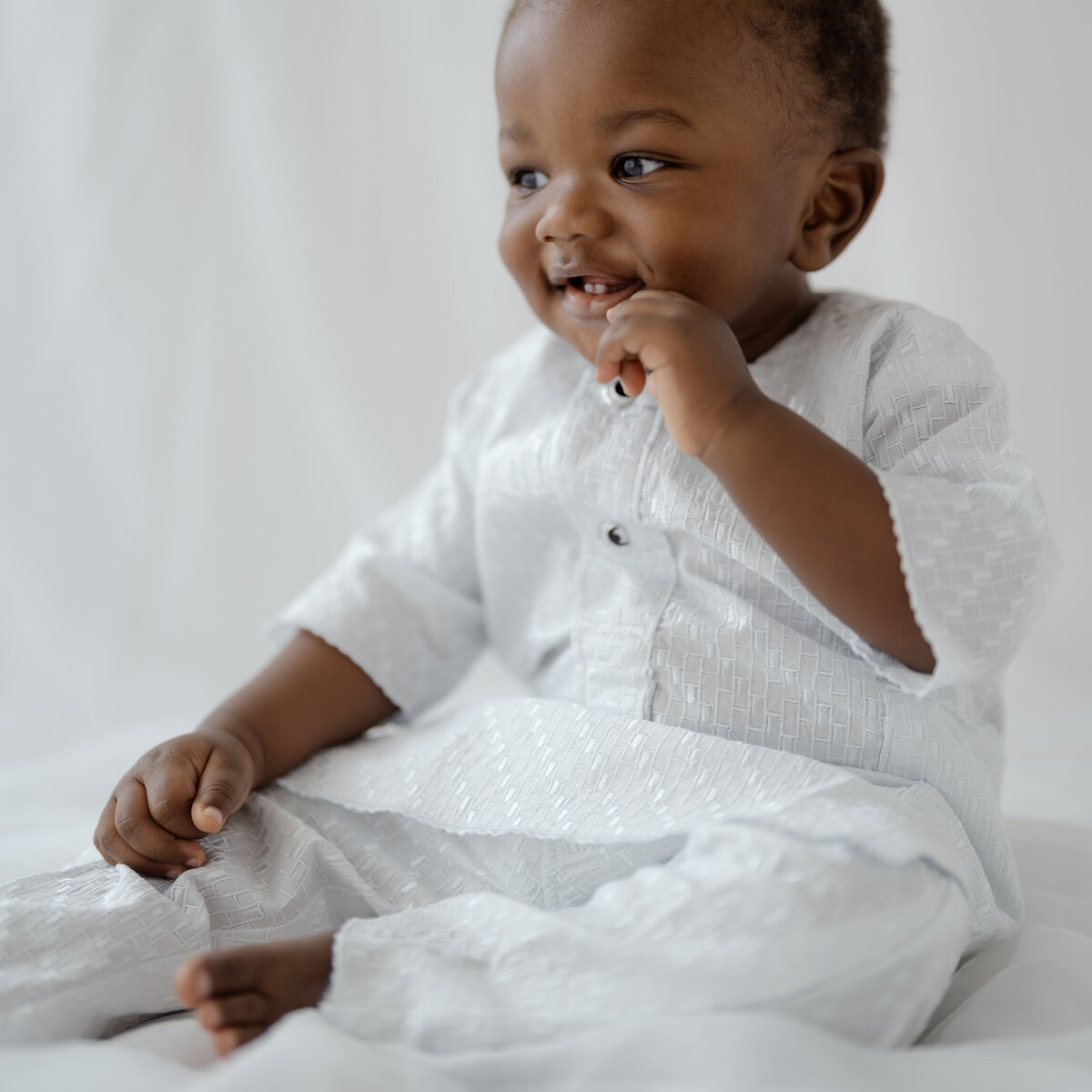 Elegant portrait of a one-year-old boy in a Fort Worth studio, captured by Nerissa James. The boy smiles sweetly while sitting in a white traditional outfit, showcasing the timeless charm and innocence of this special milestone.