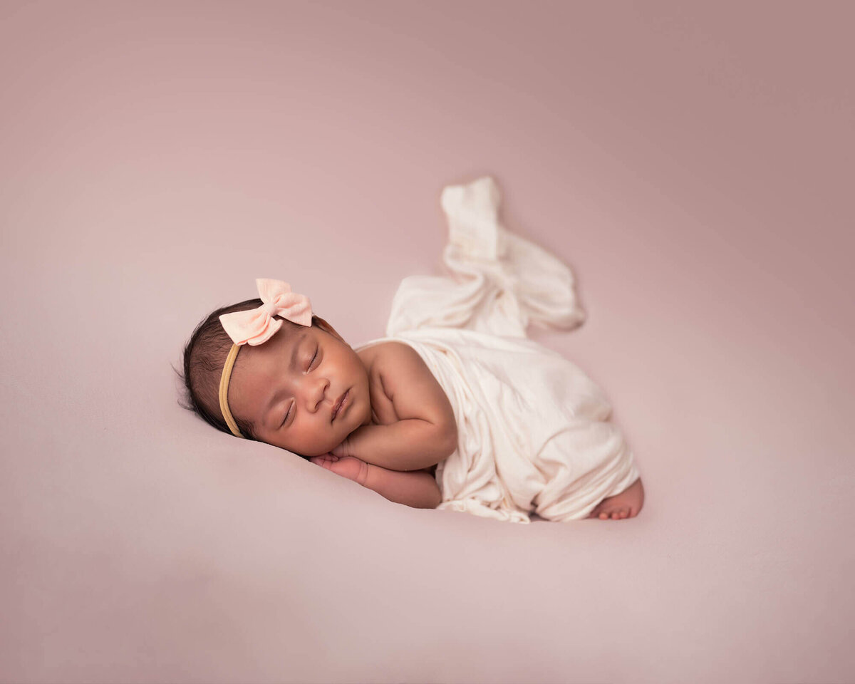 A newborn baby girl with a pink bow sleeps on a pink blanket with her hands folded under her head