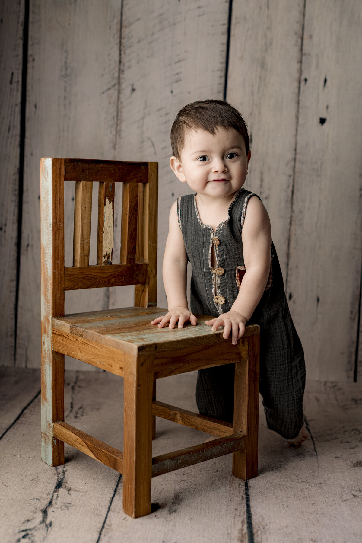 1 year old baby boy stands next to chair