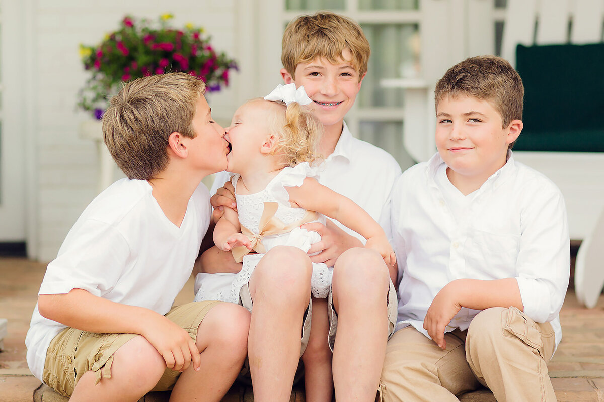 Three boys with their young sister, she is kissing one of the boys. They're all wearing white.