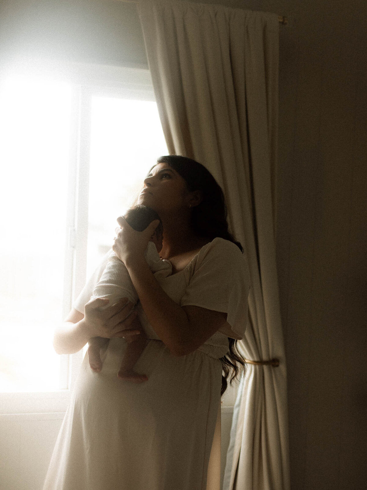 Mother in a white dress cradling her newborn by a softly lit window, intimate lifestyle newborn photography.