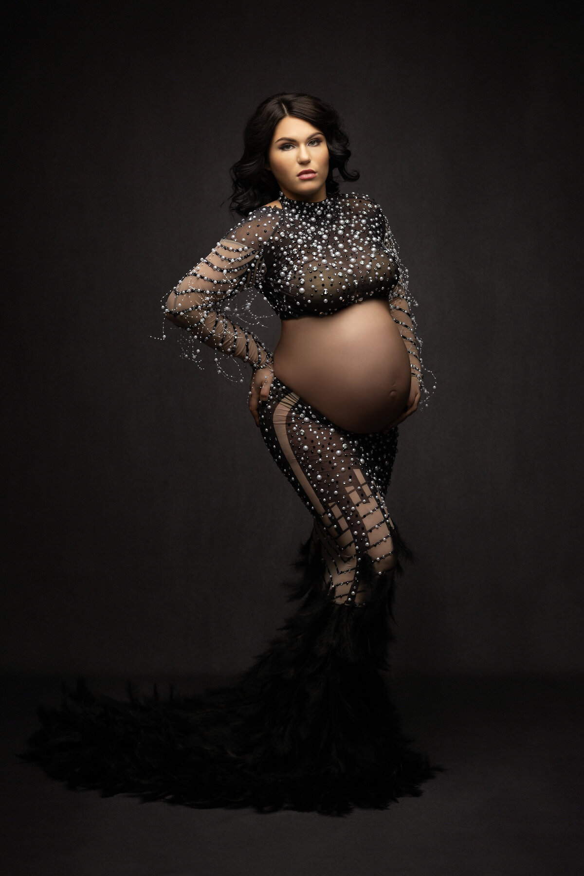 A stunning maternity portrait featuring a woman wearing a dazzling black sheer gown adorned with sparkling crystals and intricate beading. The luxurious black feathers at the hem of the gown create a dramatic, high-fashion look, captured by Lindsay Walden Photography.