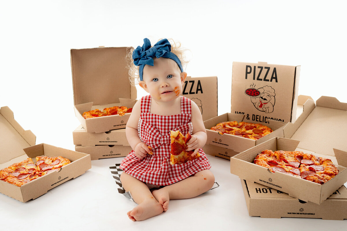 Child in a red dress surrounded by open pizza boxes, holding a slice of pizza.