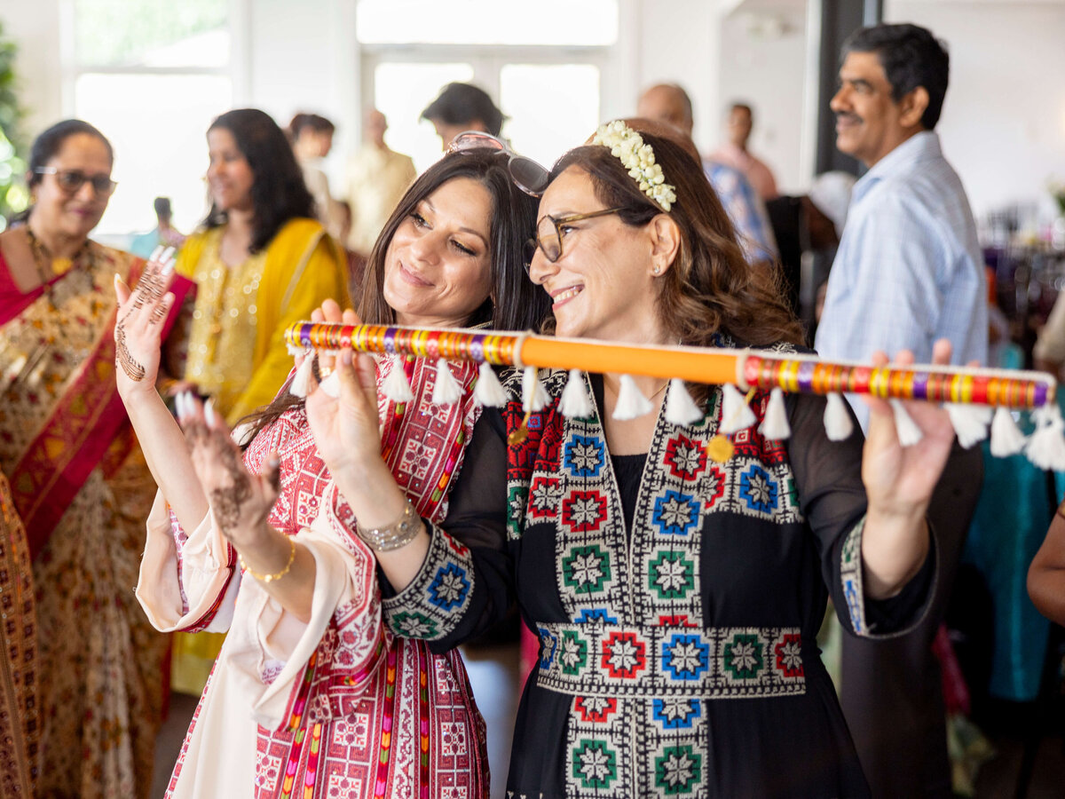 Two women in colorful traditional attire dance joyfully with a decorative stick at a lively indoor gathering. Other attendees, also dressed in traditional clothing, are seen in the background, enhancing the festive atmosphere.