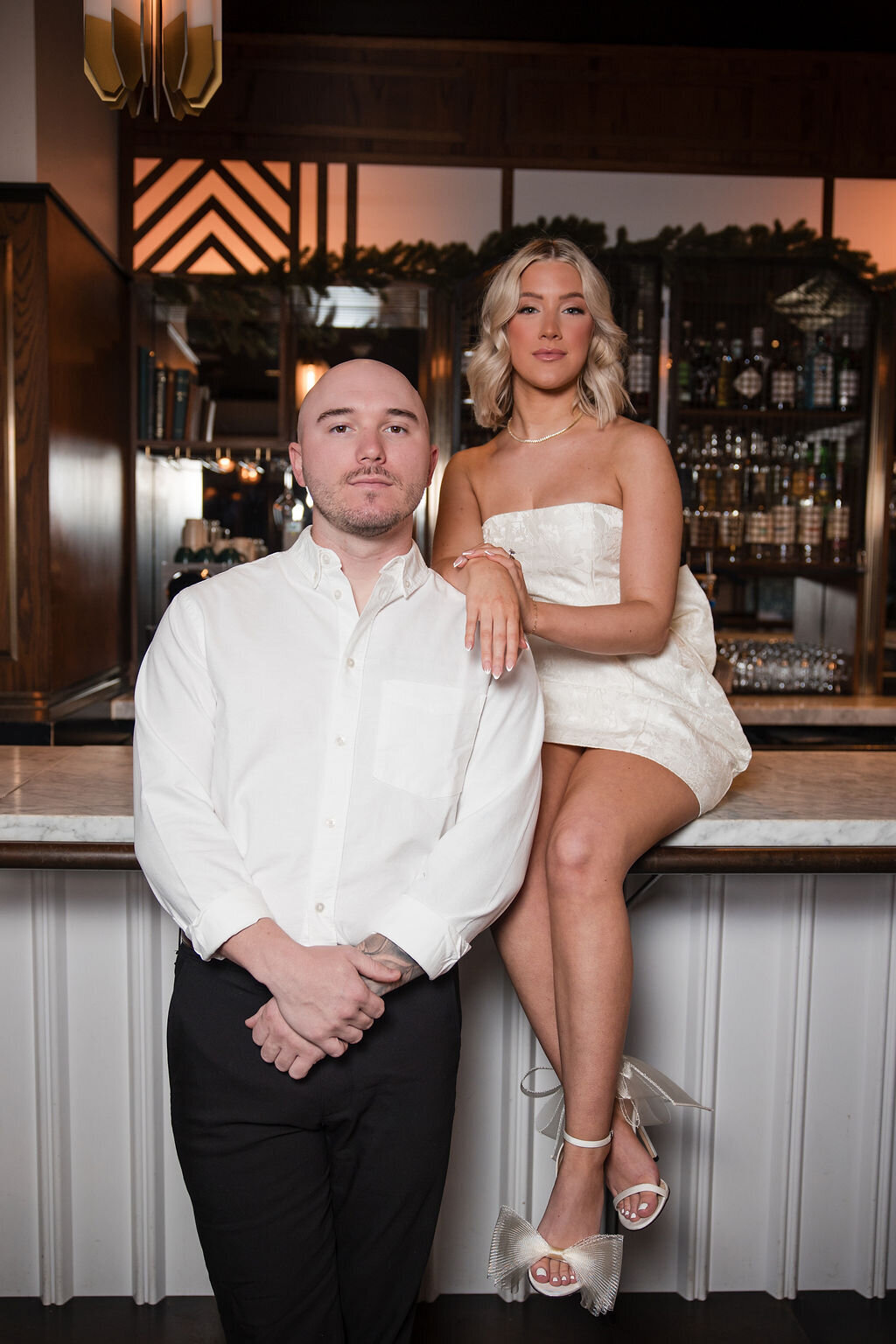 A stylish couple poses confidently at a luxury hotel bar during their engagement session. She’s seated on the counter in a chic strapless white mini dress and heels, while he stands beside her in a crisp white shirt and dark trousers. Captured in an elevated, editorial style by Phavy Photography—perfect inspiration from a luxury St. Augustine engagement photographer.
