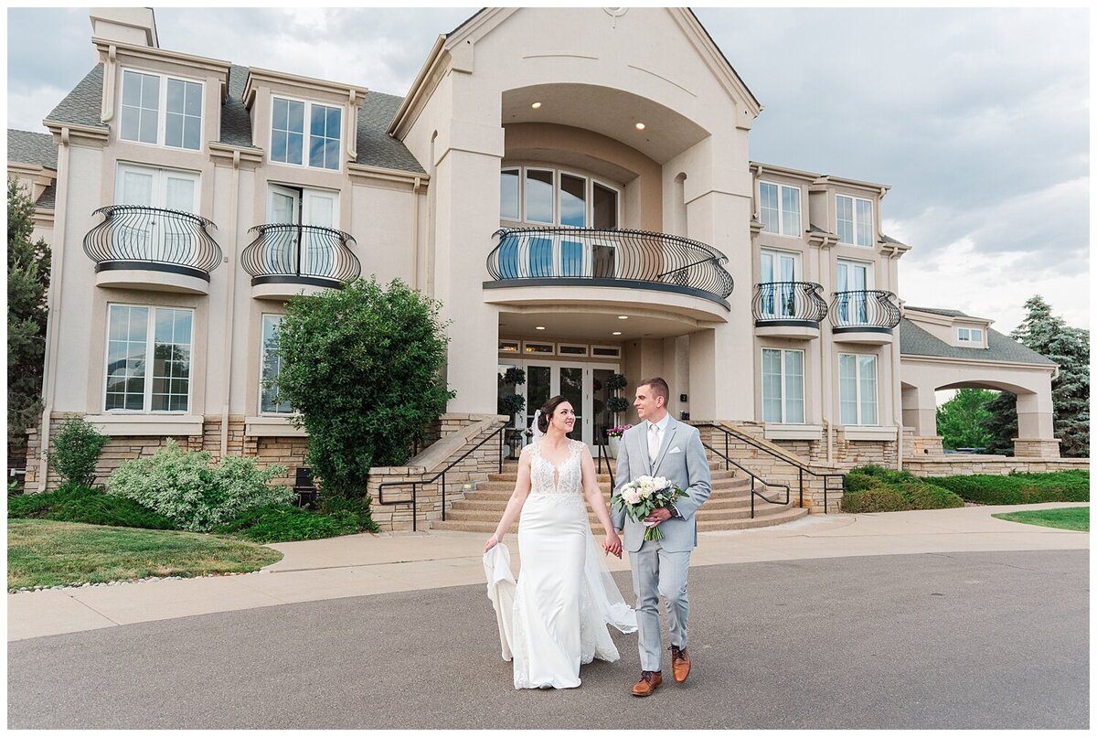 Bride and groom walking hand in hand looking at each other and walking in front of their venue The Chateaux at Fox Meadows captured by Denver Wedding Photographer