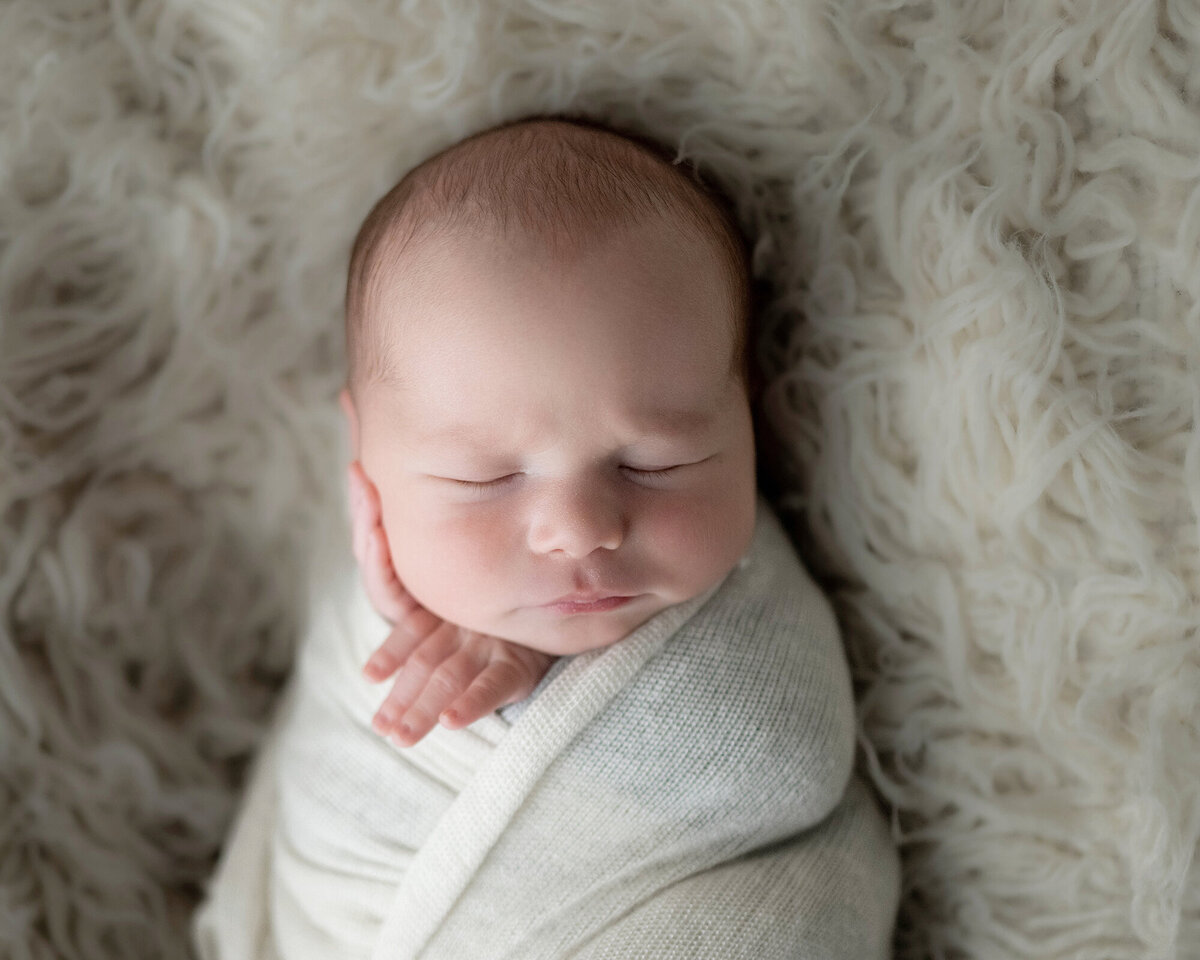 A newborn sleeps on a fur blanket in a swaddle with a hand on its cheek