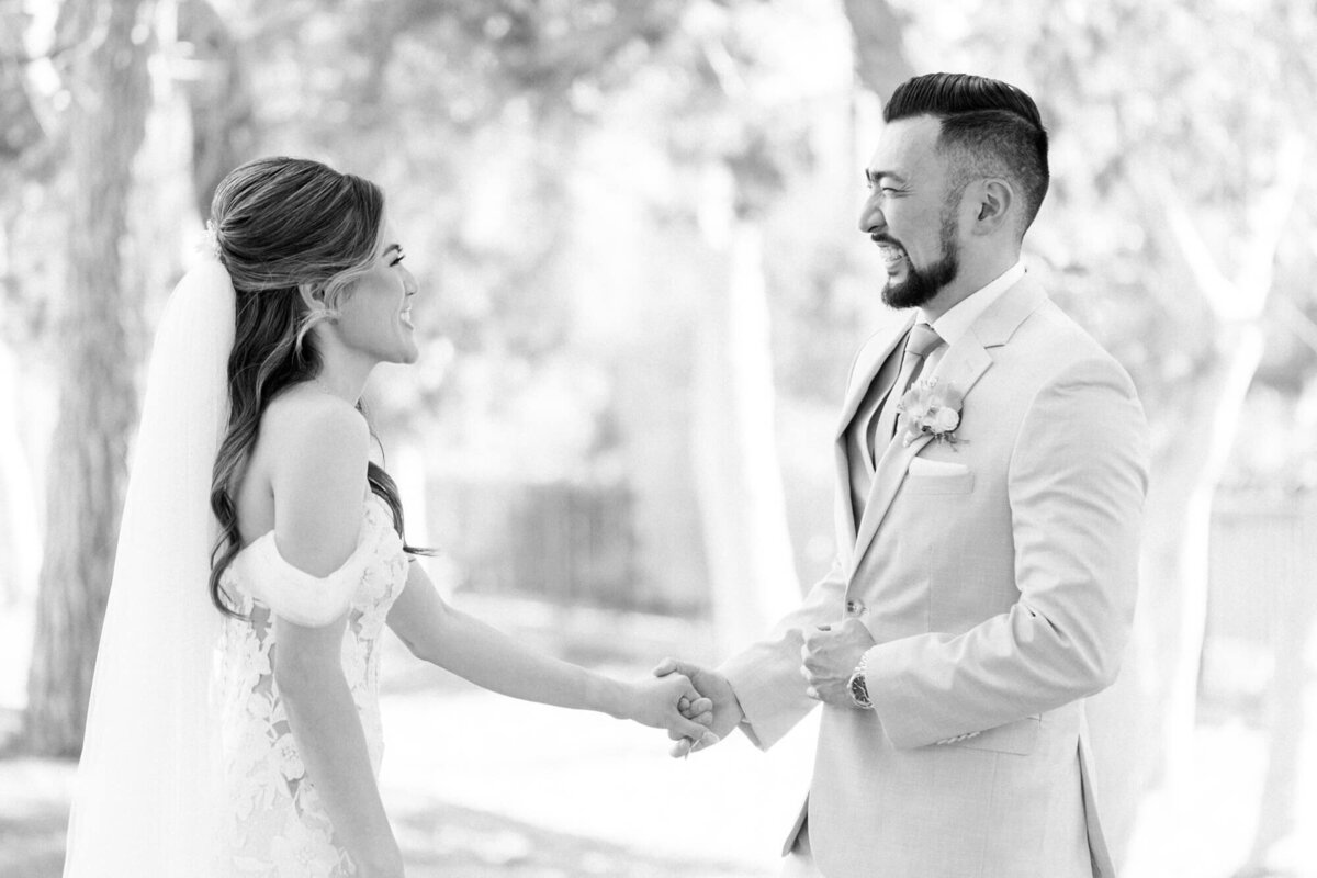 A bride and groom hold hands, smiling joyfully at each other. The bride wears an off-shoulder lace gown and veil, and the groom dons a light suit with a boutonniere. The scene is set outdoors in a softly blurred background of trees.