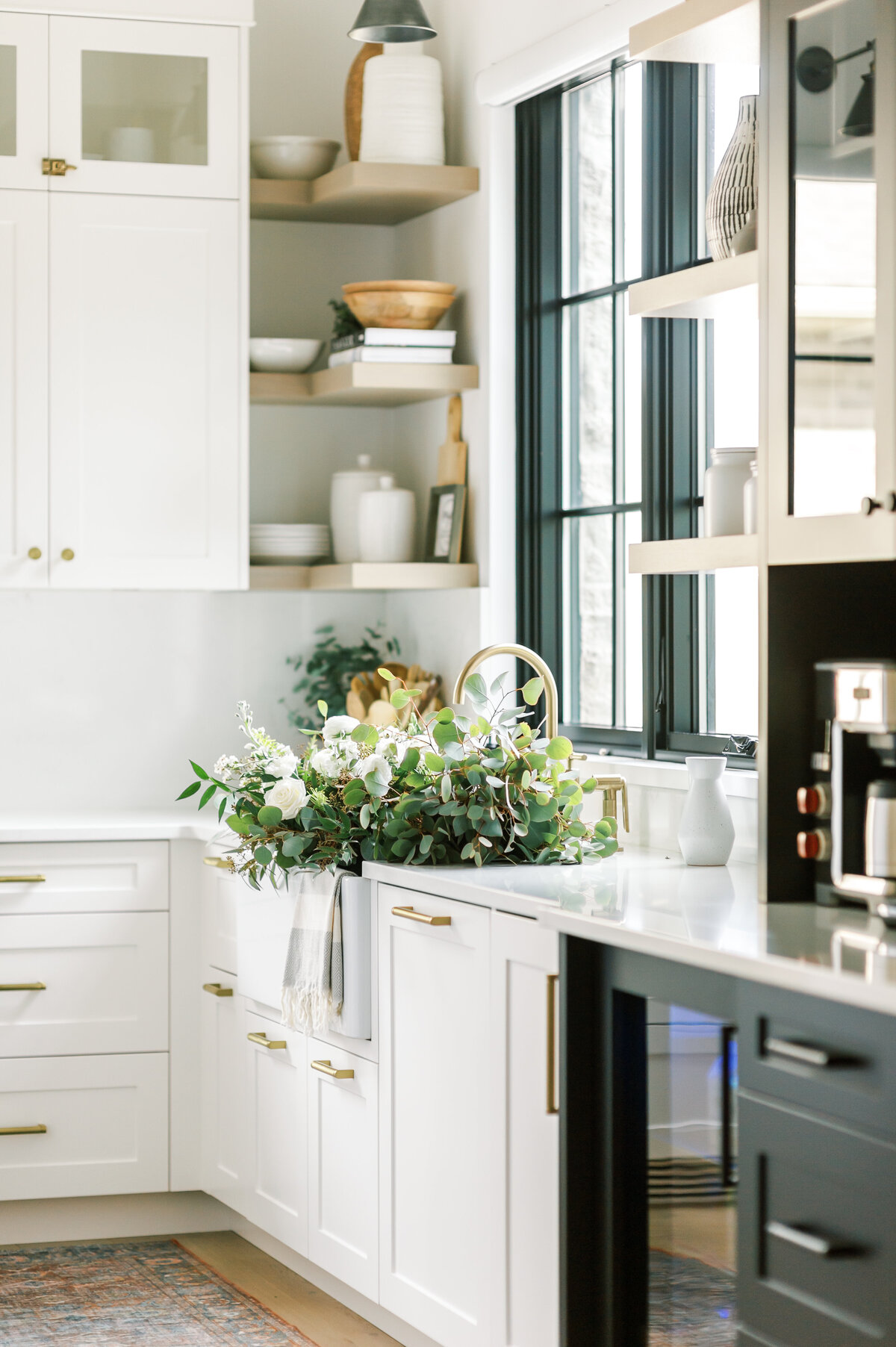 Image of a modern transitional kitchen with a mix of white shaker-style cabinetry, a black coffee bar, and natural wood tones. The farmhouse sink if full of eucalyptus and fresh-cut roses. Designed by Best London, Ontario interior designer Jen Sottosanti.