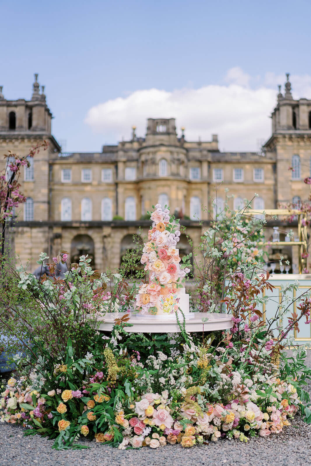 wedding cake elaborately decorated with pink and peach sugar flowers displayed in a large meadow of pink and peach flowers in front of blenheim palace