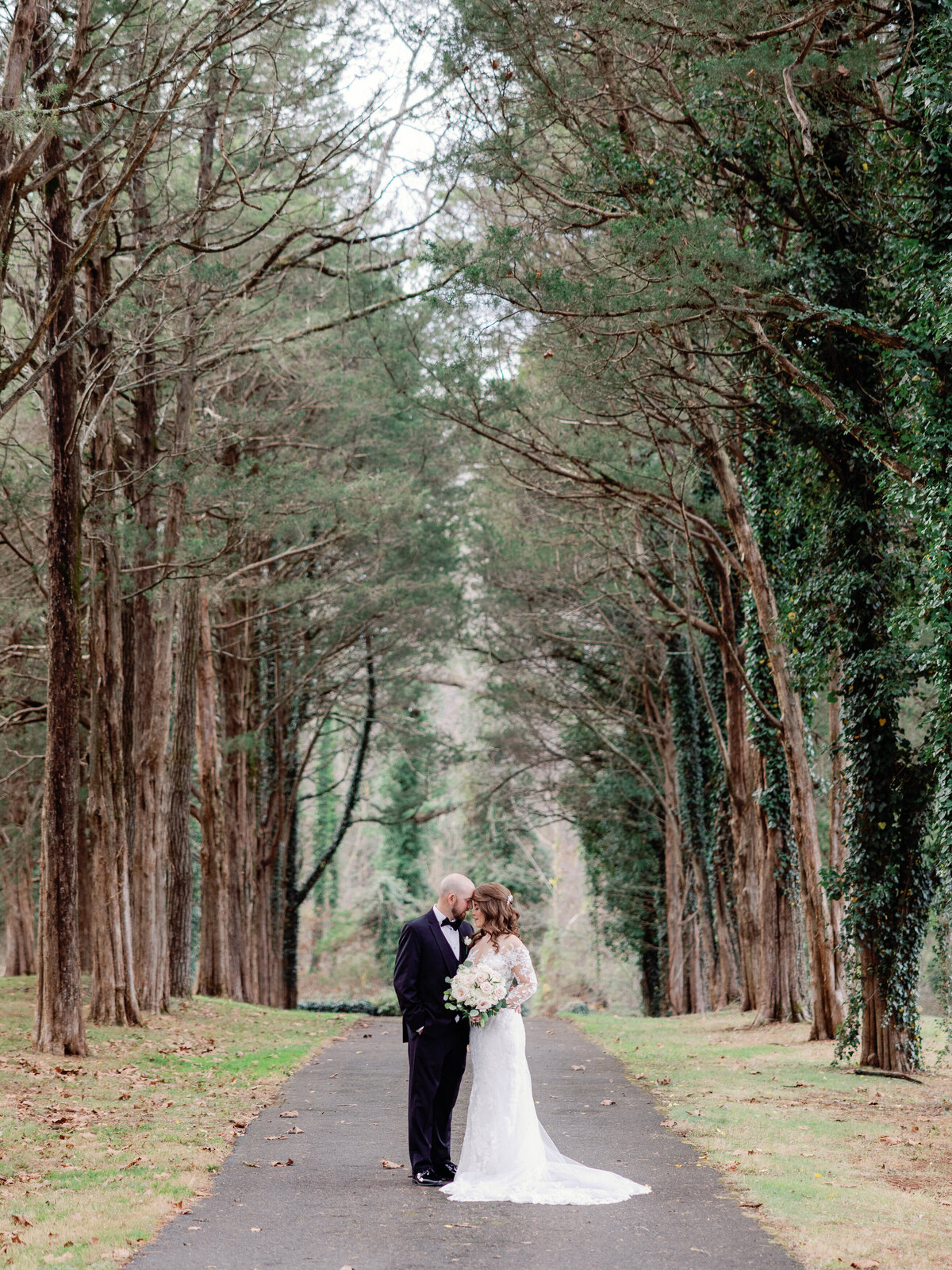 A bride and groom look lovingly at each other as they stand on a pathway underneath a parting of tall trees covered in green ivy