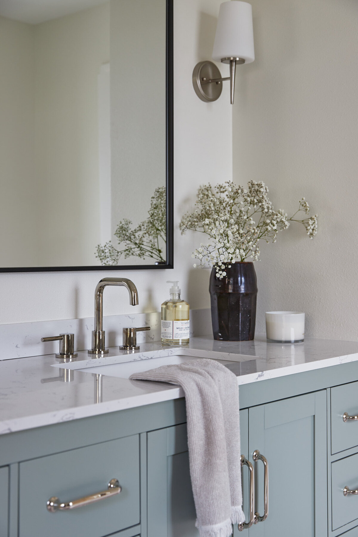 Bathroom with light blue bathroom vanity with silver cabinet pulls. A silver sink faucet with a black mirror above. Next to the sink is a dark brown vase with baby’s breath flowers, a Handsoap pump, and a white candle. Next to the vanity is a white wall with recessed white oak shelves with photos, jars, and a woven basket.