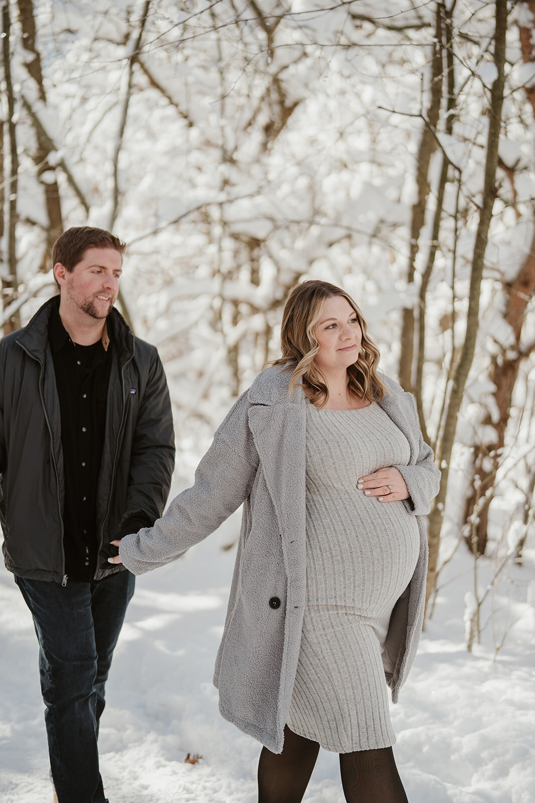 Expecting couple holding hands while walking through a snow-covered trail at Al Sabo Preserve during their winter maternity photography session.