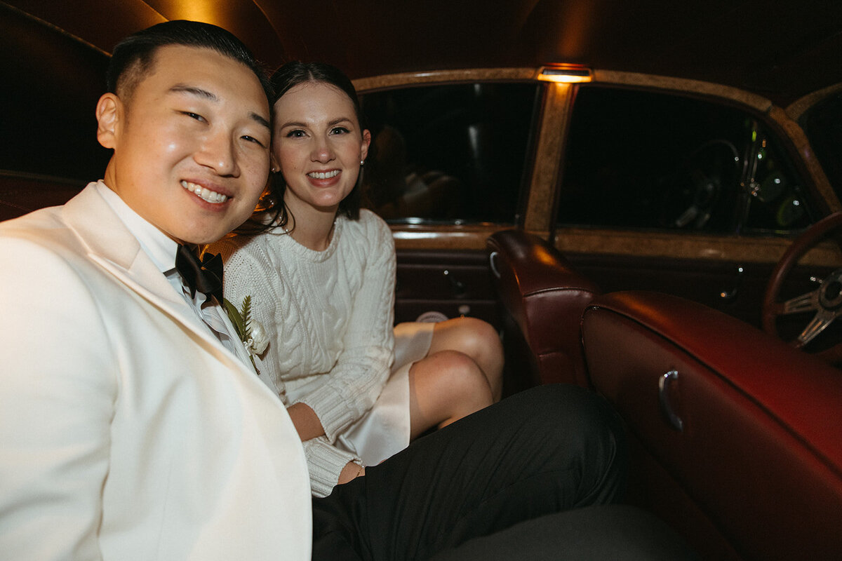 Bride cozying up to groom with joyful smiles during sendoff in old historic car. 
