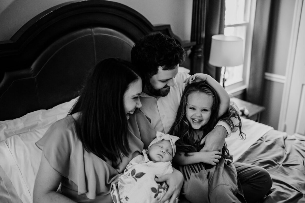 Family photographers Maryland captures in home photo session with parents sitting on their bed with their young daughter and newborn baby while they laugh together