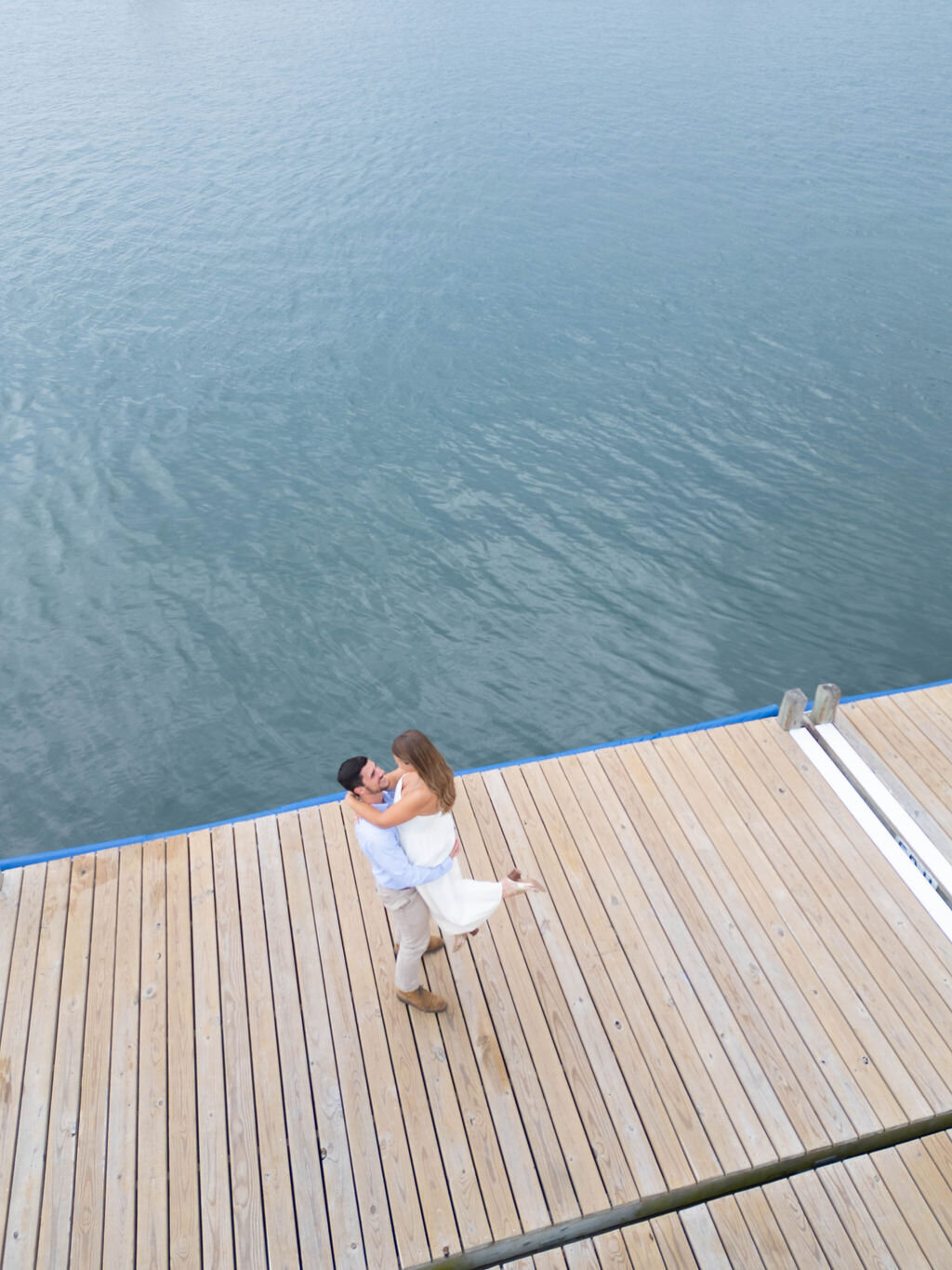 Wedding Photographer RI | A couple embraces joyfully on a wooden dock overlooking calm water. The man lifts the woman, capturing a moment of happiness and romance.