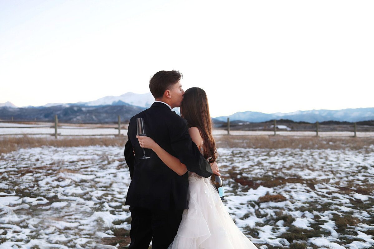 The bride and groom stand facing the mountains, holding champagne, hugging in a beautiful snowy scene,