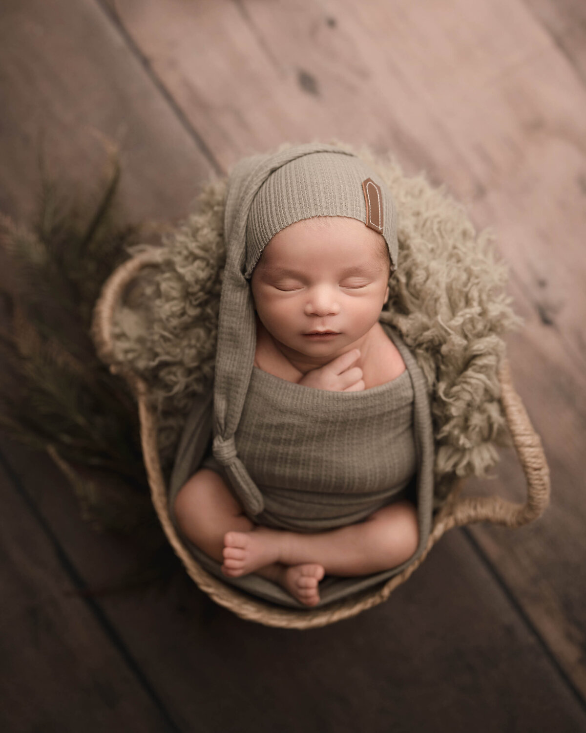 Aerial image of newborn photoshoot. Baby boy wrapped in taupe has his legs folded atop of him as he is posed in a basket. His fingers are peeking out of the wrap and he is wearing a long cap. Baby boy is sleeping peacefully. Captured by best Murrieta newborn photographer Bonny Lynn Photography.