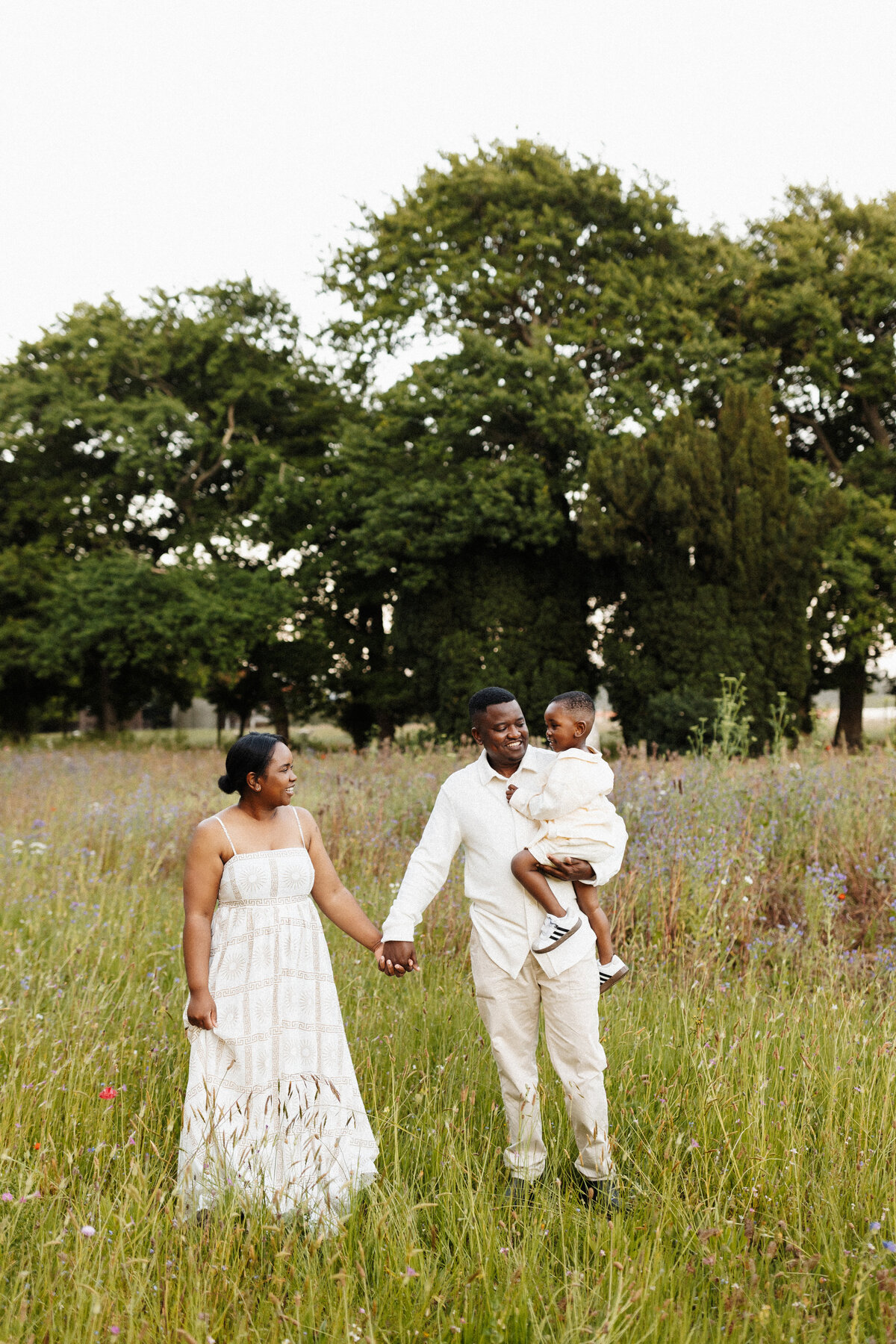 A mother and father holding hands while the father carries his little boy during the photoshoot in a field of grass in christchurch with family photographer lucy jane