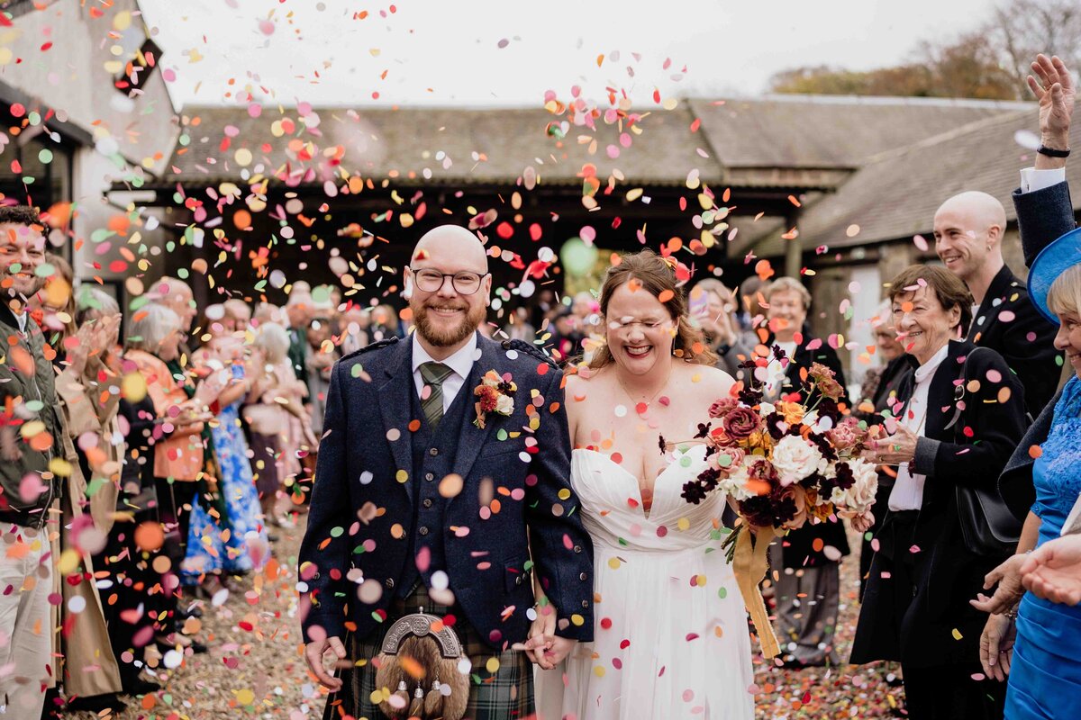 A joyful wedding scene shows the bride and groom smiling as they walk through a shower of colourful paper confetti. The groom wears a dark suit with a kilt, and the bride wears a white gown and holds a bouquet. They are photographed surrounded by celebrating guests outdoors under the Aberdeenshire sky.