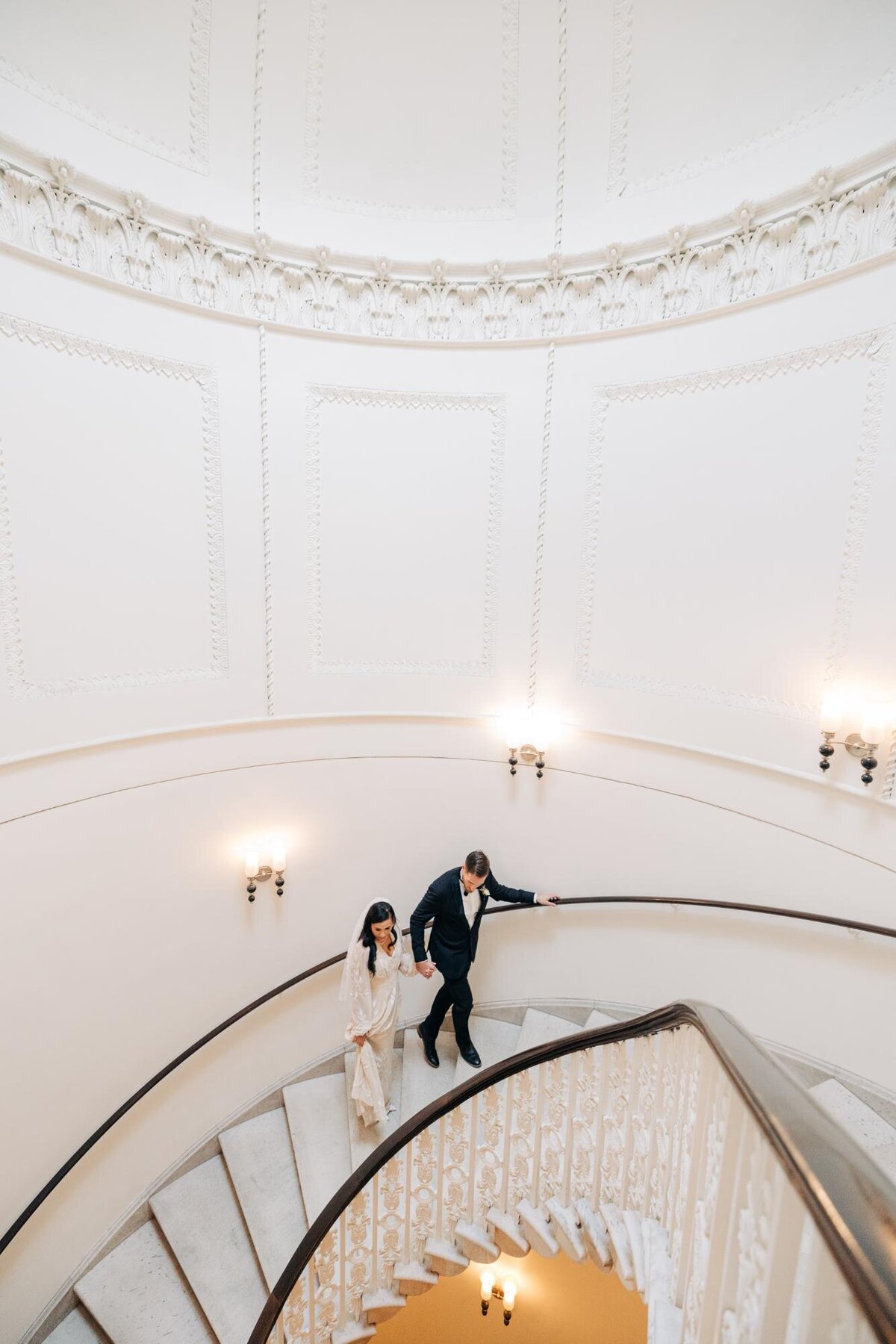 A couple holding hands while descending a grand spiral staircase in an elegant interior.