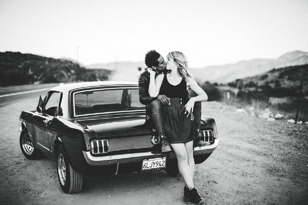 Bride to be leans back against her fiance while he sits on the trunk of his Mustang and they share a kiss
