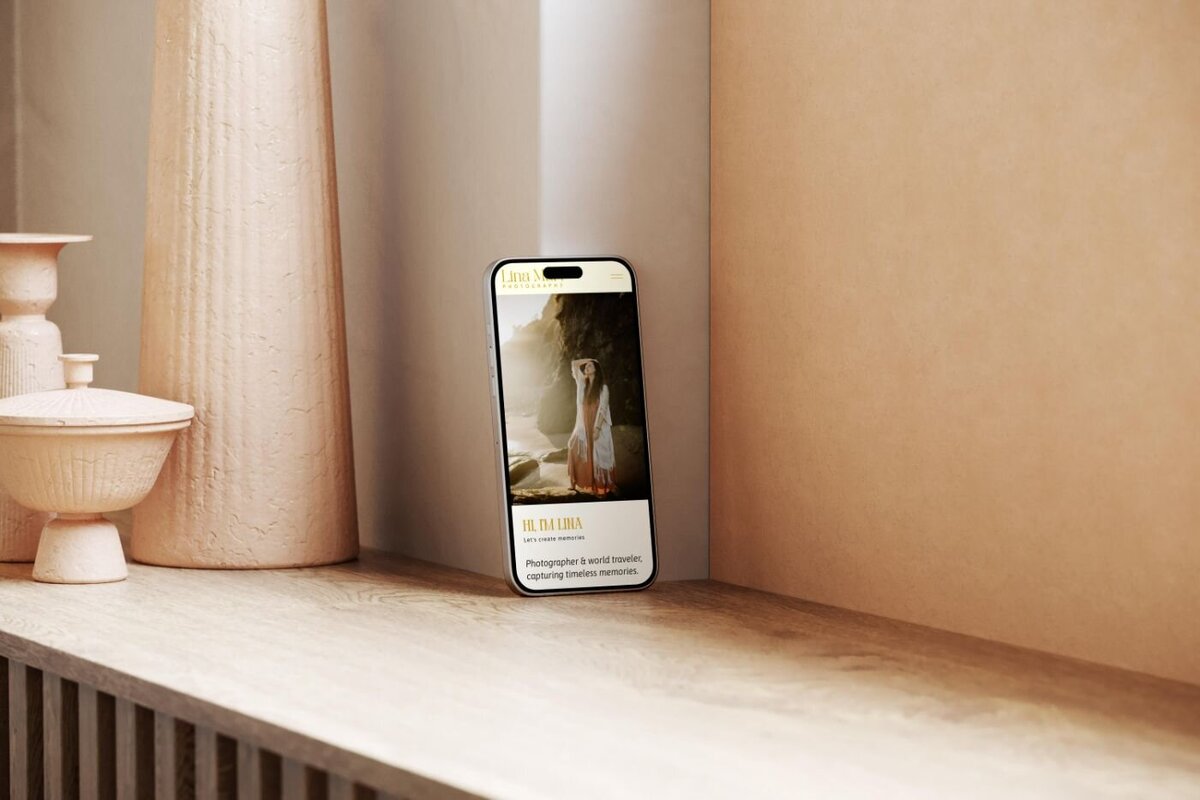 Smartphone propped against a wooden shelf and ceramic vases, displaying the Lina Mart Photography mobile website with a full-screen beach portrait of a woman in a long dress.