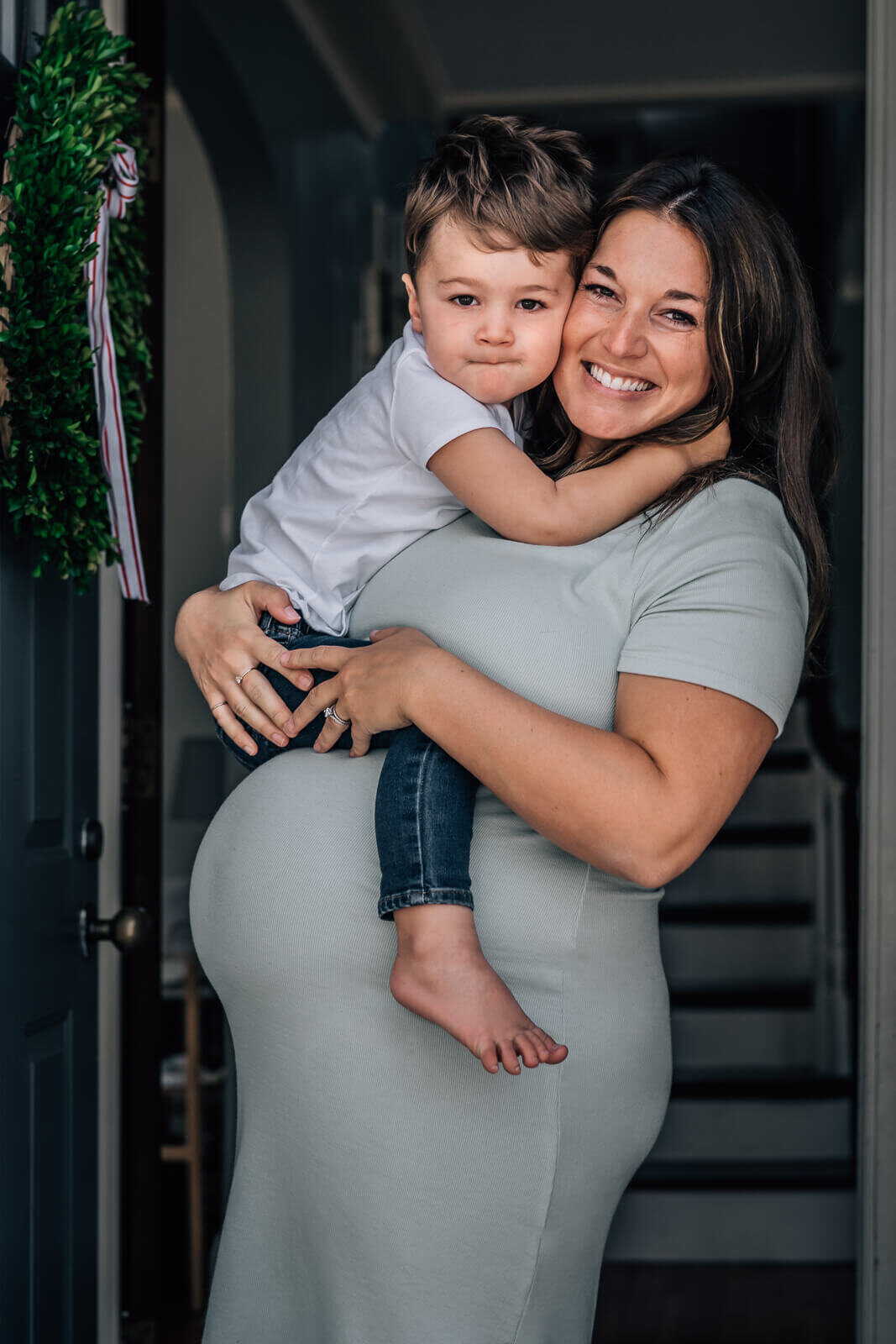 A toddler straddles his mother, atop her baby bump, framed by a doorway during a photo session.