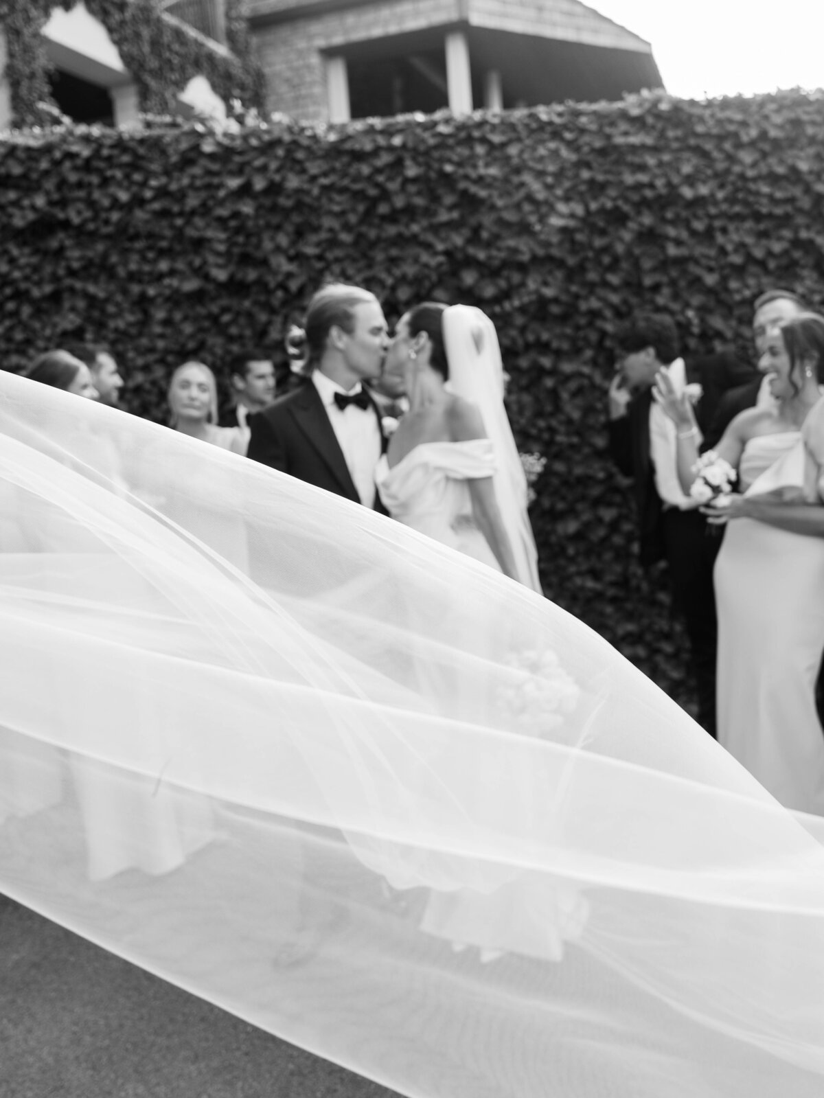black and white photo  of wedding party cheering while the bride and groom kiss and the veil swoops to the foreground of the photo taken by Mansfield Wedding Photographer, Annie Trammel