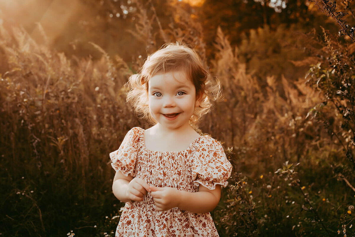 a little red headed girl pinching her fingers together smiling at the camera in the sunset