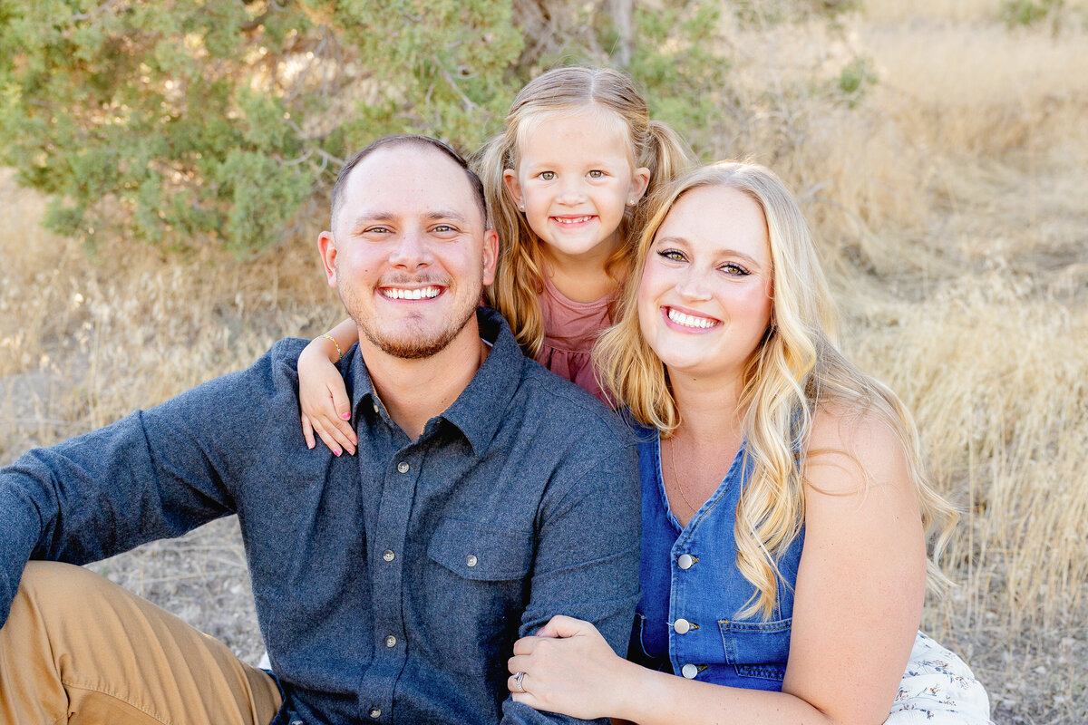 A close-up portrait of a happy family sitting together in the golden fields of Eagle Mountain, Utah. The daughter leans on her father’s shoulder, and all three are beaming at the camera