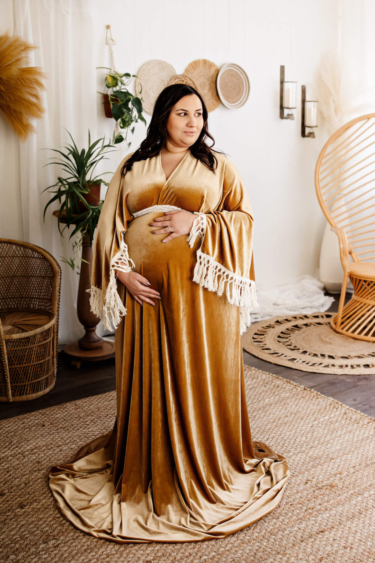 A pregnant woman in a long, gold velvet gown with fringed sleeves stands indoors, resting her hands on her belly. Surrounded by wicker furniture, plants, and neutral boho-style decor, this scene is perfect for stunning Bloomington maternity photographer sessions.