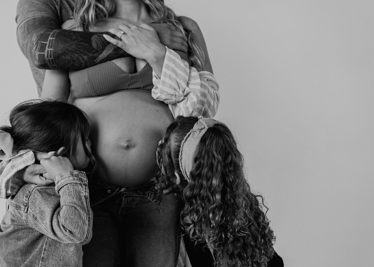 black and white close-up image of pregnant belly with husbands tattoos and 2 little girls kissing the baby bump