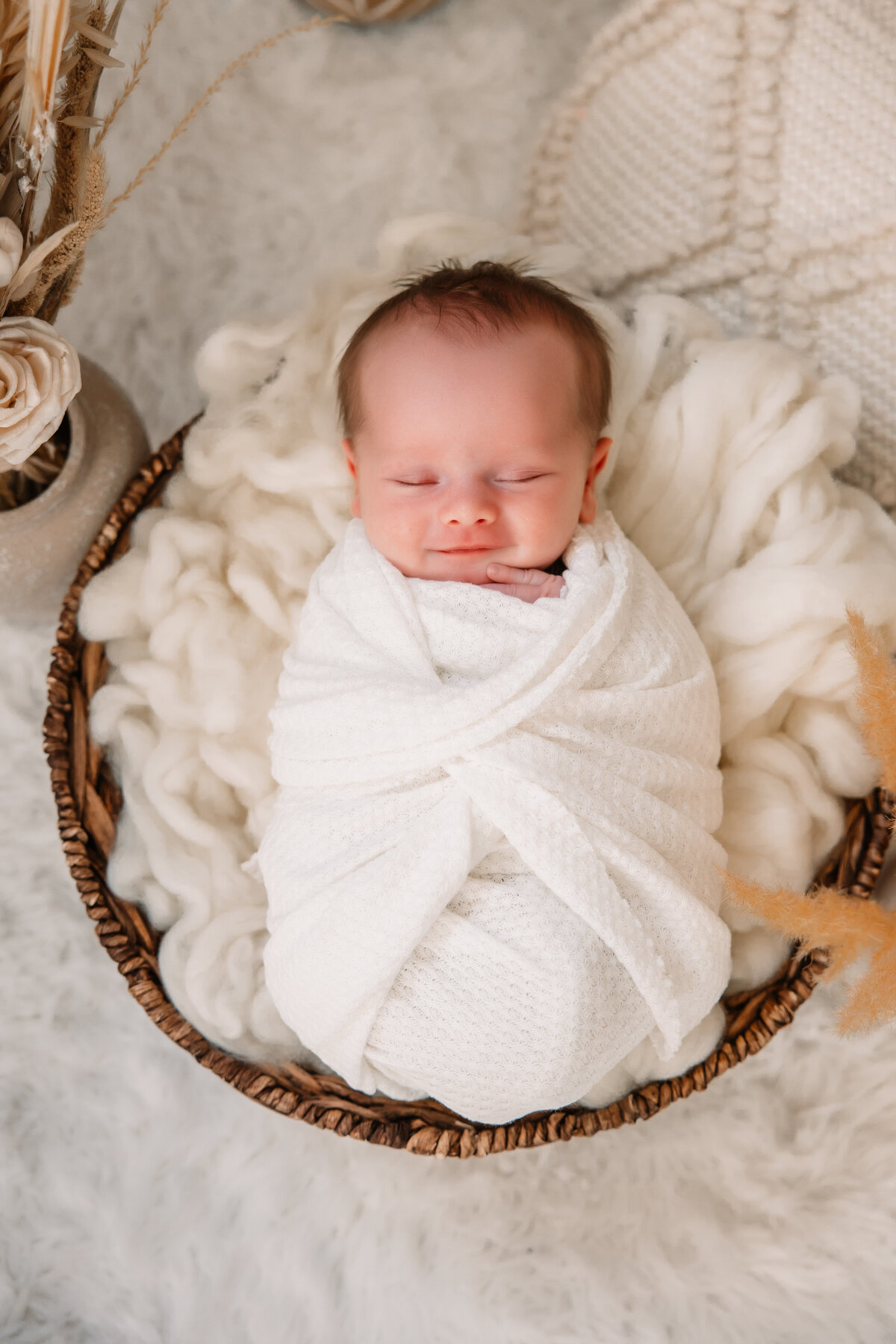 A smiling newborn baby in a wicker basket wrapped in white swaddle