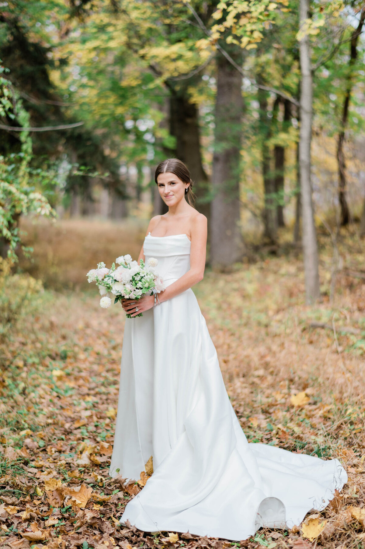 The bride poses gracefully in a forest setting, holding a lush bouquet of white and blush flowers