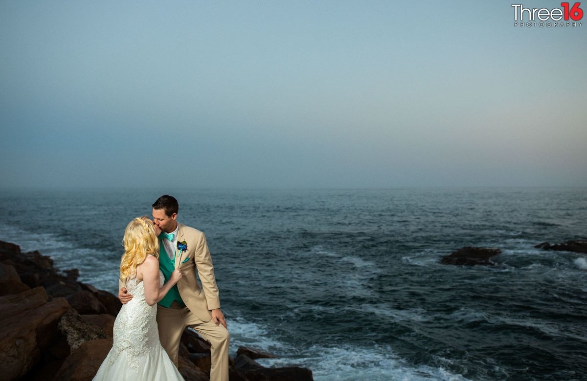 Bride and Groom share a kiss while standing on the rocks of the beach