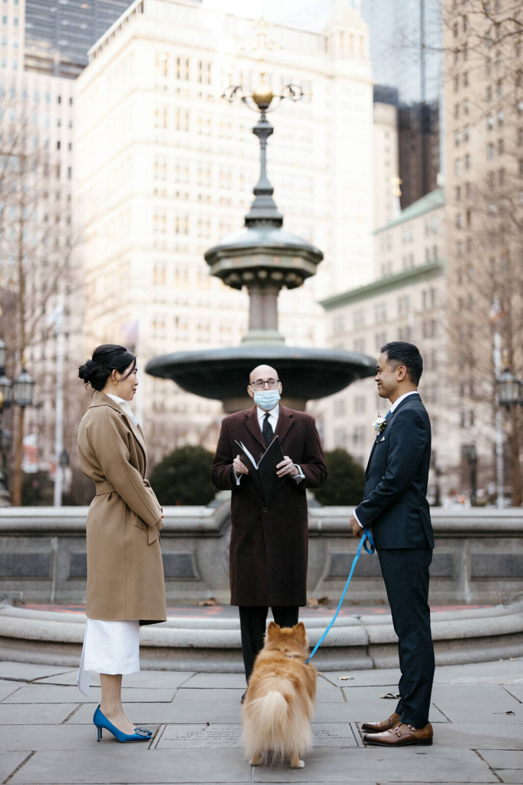 The bride and the groom, with their dog, saying their vows to a wedding officiator in front of NYC City Hall Park fountain.