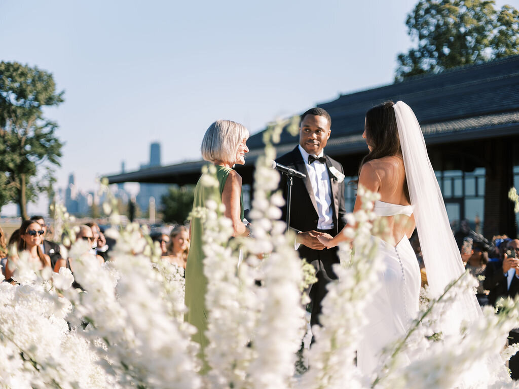 The bride and groom hold hands while exchanging vows, with a city skyline and floral décor framing the moment.