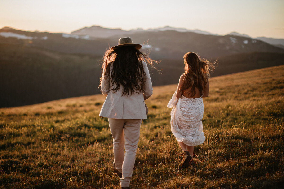 Groom wearing an ivory wedding suit and hat walks with bride wearing a white wedding gown on a field during golden hour.