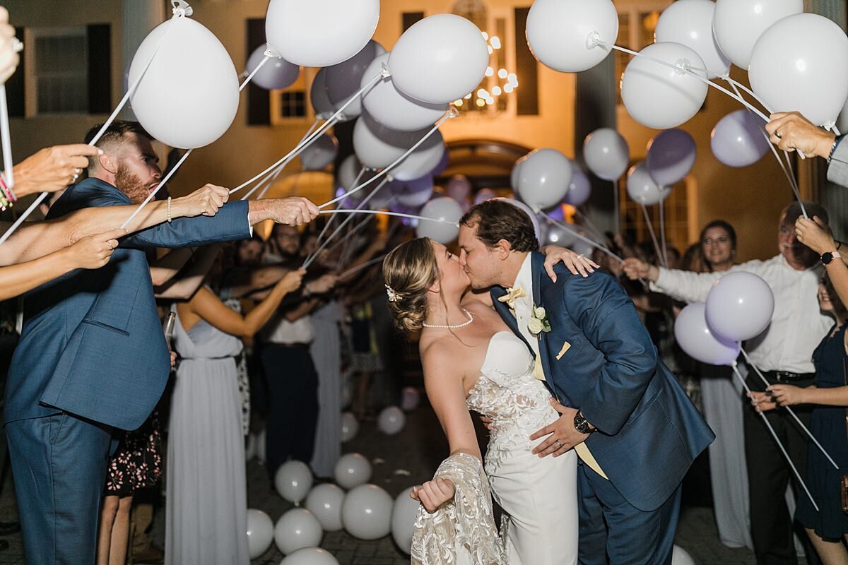 The groom, wearing a dark blue suit dips the bride, wearing a strapless wedding dress, into a kiss as their guests wave balloons on sticks and cheer as they leave their wedding at The Estate at Cherokee Dock.