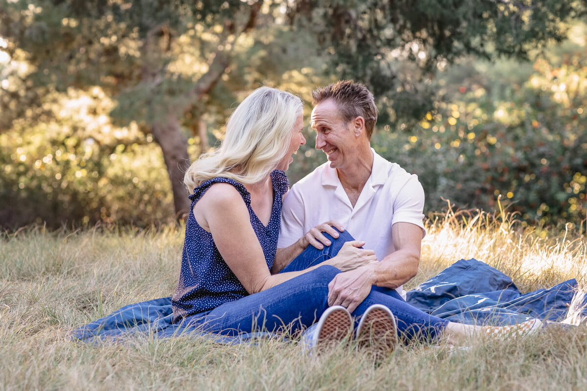 A husband and wife leaning close to each other and laughing, sitting on a dark blue picnic blanket in the grass, surrounded by greenery at Jeffrey Open Space.