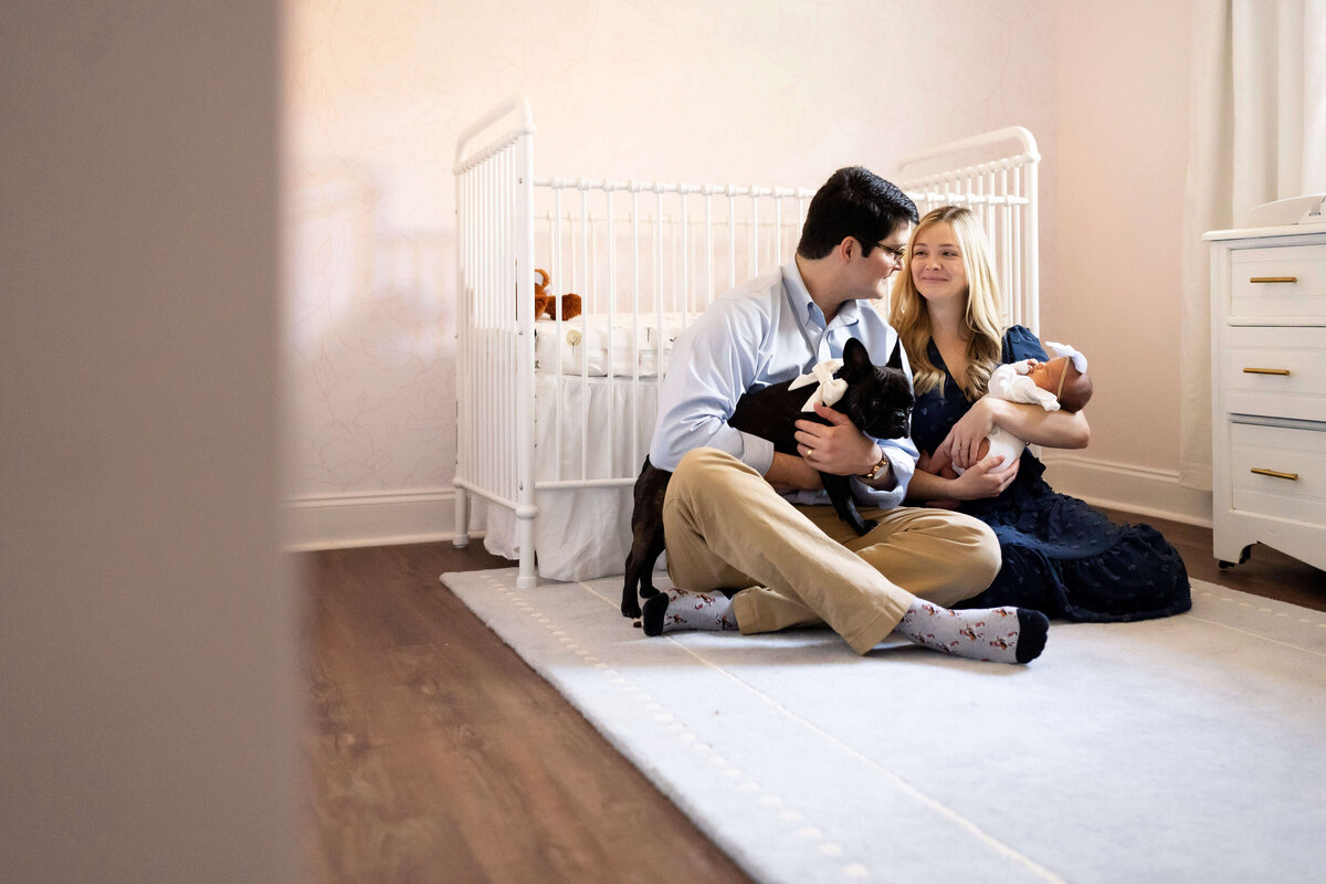 Parents sitting on the floor in front of crib looking at each other while holding baby and dog