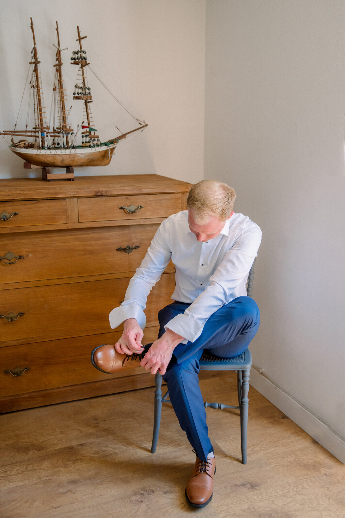 Destination Wedding in Spain | A groom in blue dress pants and a white button-up shirt sits in a wooden chair to tie his shoes. Behind him is a wooden dresser with a model sailboat on top. Captured in Spain by luxury film wedding photographer, Caitlin Wilcox.