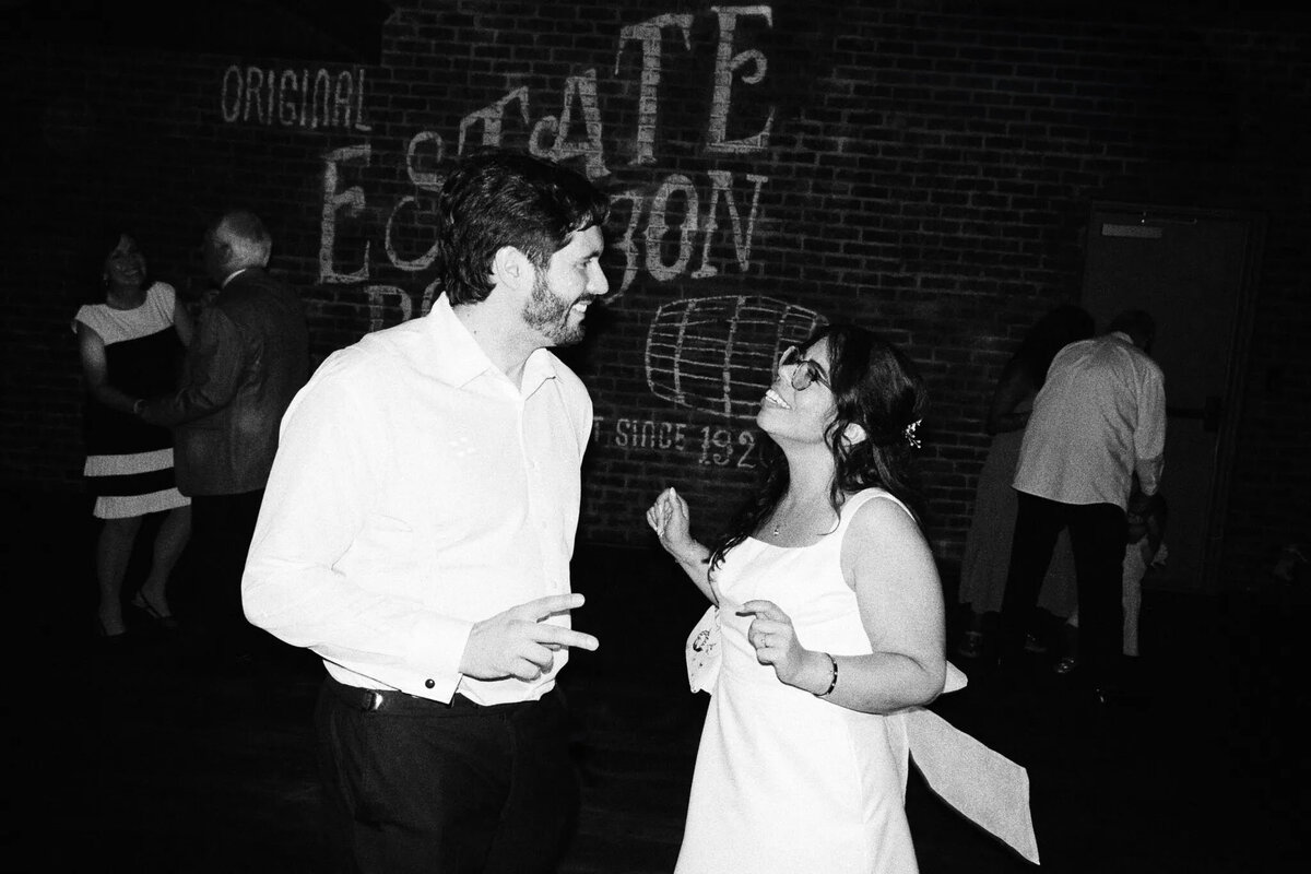A man and a woman, both dressed in white, smile and dance together in front of a brick wall at a party. Captured by an NJ wedding photographer, the black and white photo shows other guests mingling and dancing in the background.